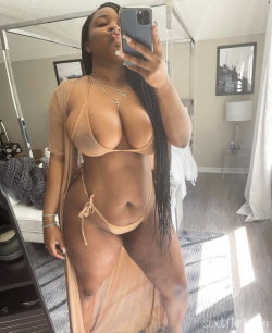 A curvy woman with long dark hair takes a mirror selfie in a beige bikini and sheer cover-up, posing in a well-lit