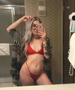 A woman with long silver hair and glasses takes a mirror selfie in a red bikini, wearing a patterned robe open over her