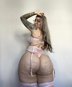 Curvy woman with long blonde hair and full tattooed arm poses in pink lace lingerie against a plain wall.