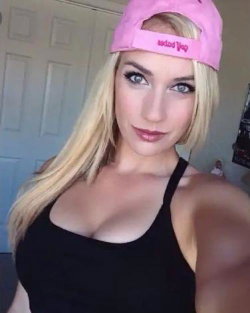 Blonde woman in a pink cap and black tank top takes a selfie indoors, showing a curvy figure with defined shoulders.