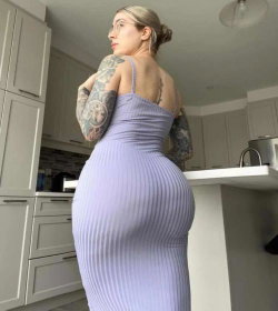 A woman with blonde hair and extensive arm tattoos wears a tight purple ribbed dress, standing in a kitchen with her