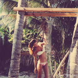 Curvy woman in white bikini stands under outdoor shower surrounded by palm trees, holding showerhead with one hand.