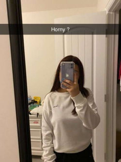 Woman with long dark hair takes mirror selfie in white sweatshirt and black bottoms, holding phone to face.