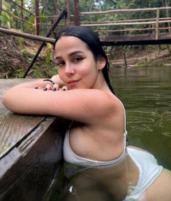 Brunette woman with wet hair in a white bikini top, leaning on a wooden dock in a natural water setting.