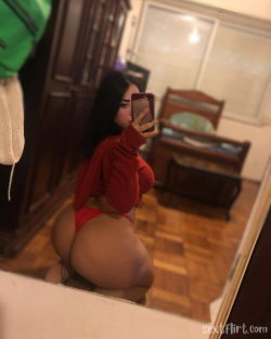 Curvy woman with long dark hair takes mirror selfie in red top and red thong, kneeling in bedroom with wooden furniture.