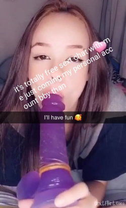 Brunette woman with long hair in a dimly lit room, holding a purple sex toy close to her mouth while looking at the