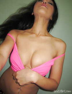 Brunette woman with curvy figure wearing a pink top, posing with head tilted back and hand near chest.