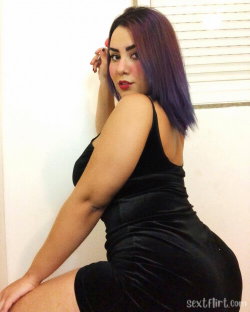 Curvy woman with purple hair in a black velvet dress posing indoors, hand near face.