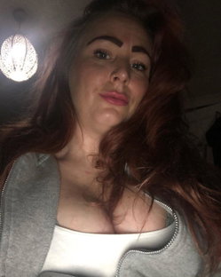 Redheaded woman with long wavy hair, wearing a gray zip hoodie over a white top, taking a selfie under a decorative