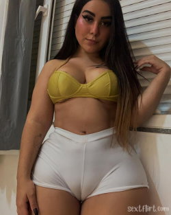 A curvy woman with long dark hair wears a yellow bra top and white high-waisted shorts, posing indoors near a window