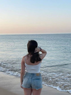 Woman with dark hair in a white tank top and denim shorts stands on a beach facing the ocean at sunset, hand in her