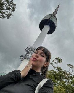 A young woman with dark hair and bangs poses in front of a tall observation tower, wearing a black long-sleeve top and