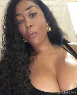 A woman with long curly hair and dark skin poses in a black top, looking at the camera with lips puckered.
