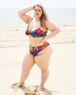 Curvy woman in colorful bikini posing on a sandy beach, hand on hip and head, under bright sunlight.