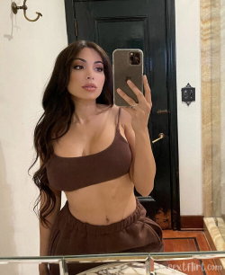 Brunette woman with long wavy hair takes mirror selfie in brown crop top and matching pants, standing indoors near a