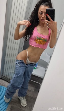 Brunette woman in a pink crop top and low-slung jeans takes a mirror selfie, showing a slim build and lace underwear.