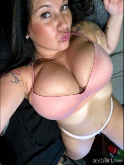Brunette woman with tattoos wearing a pink bra and white thong, lying on her side in a close-up selfie pose.