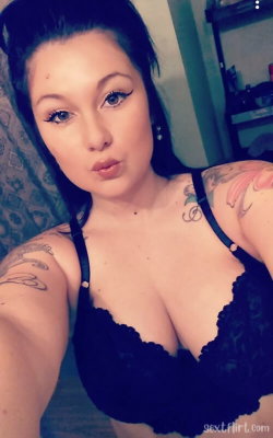 A woman with dark hair and tattoos on her arms and shoulder wears a black lace bra, posing in a selfie-style shot