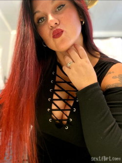 Woman with long red hair and dark makeup poses in a black lace-up top, hand on neck, indoors.