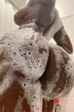 A person with dark skin is covered in soap suds while bending over in a shower, back and buttocks visible.