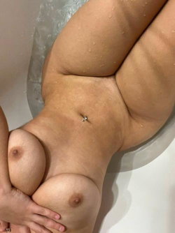 Curvy woman with bare breasts and a belly piercing lying in a bubble bath, hands covering her chest.