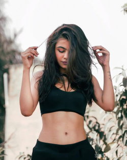 A woman with long dark hair in a black crop top and high-waisted bottoms, standing outdoors with plants behind her,