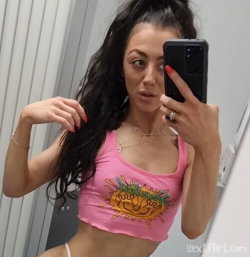 Brunette woman in a pink crop top takes a mirror selfie, showing midriff and styled hair, indoors.