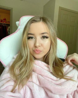 Blonde woman with long hair wrapped in a pink blanket, sitting in a white and pink gaming chair indoors.