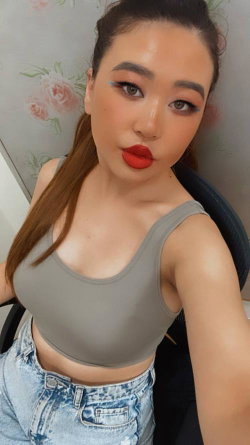 A woman with long brown hair in a high ponytail, wearing a gray crop top and light blue jeans, poses for a selfie