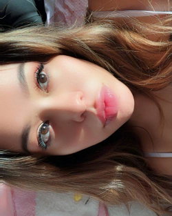 A woman with long brown hair and defined eyes lies on her side, making eye contact in a close-up selfie shot.