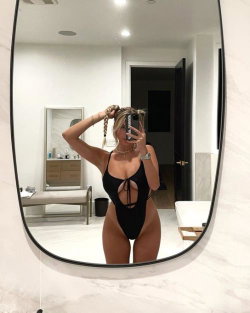 A curvy woman with blonde hair takes a mirror selfie in a black one-piece swimsuit with a front cutout, standing in a