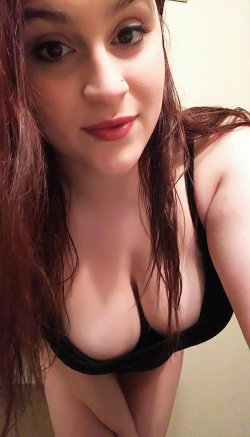 A woman with long dark hair and red lipstick wears a black bra, posing closely in an indoor setting.