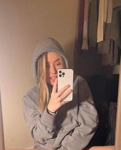 Blonde woman in a gray hoodie takes a mirror selfie, holding a white phone, in a dimly lit room with clothes visible in