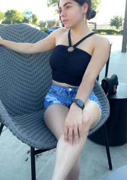 A young woman with dark hair sits on a gray chair, wearing a black crop top and denim shorts.