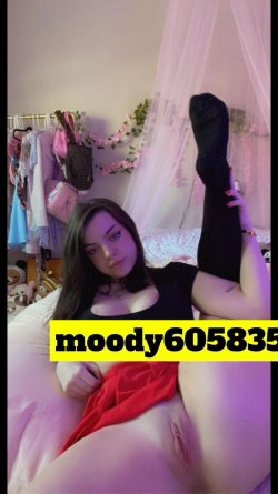 A brunette woman with a curvy build lies on a bed, wearing a black top and red skirt, lifting one leg in black