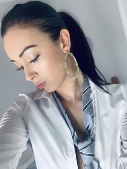 A woman with dark hair in a ponytail wears a white shirt over a striped top, posing indoors with large hoop earrings.