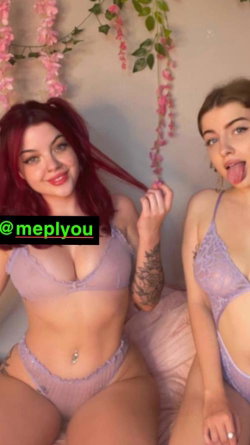 Two women in matching lavender lingerie pose together under pink floral decorations, one with red hair and a visible