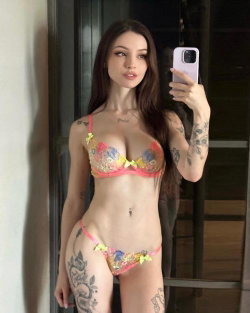 Brunette woman with tattoos in colorful lingerie takes a mirror selfie indoors, showing curvy figure and multiple