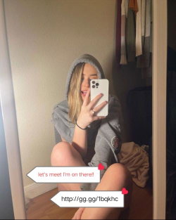 A blonde woman in a gray hoodie takes a mirror selfie, sitting bare-legged on the floor with her phone in hand.