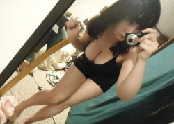 A woman with dark hair in a black top and shorts takes a mirror selfie in a bedroom, legs extended and holding a camera.