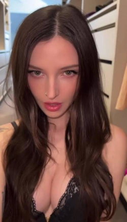 Brunette woman with long wavy hair, wearing black lace lingerie, taking a selfie in a bedroom setting.