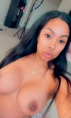 A woman with long dark hair takes a close-up selfie, bare-chested, wearing a delicate necklace in a room with shelves
