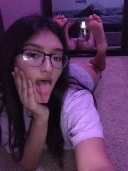 A young woman with long dark hair and glasses lies on the floor, sticking her tongue out and holding her feet up,