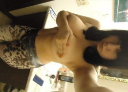 A woman with dark hair takes a selfie, partially covering her bare chest with one hand while wearing patterned pants in