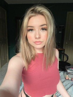 Blonde woman in a red crop top takes a selfie in a bedroom, showing a slim build and styled hair.