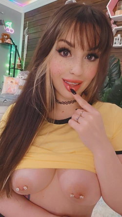 Brunette woman with freckles and piercings in a yellow crop top, posing indoors with visible nipple jewelry.