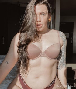 A woman with long brown hair and visible tattoos on her arms wears a beige bra and Calvin Klein underwear, posing