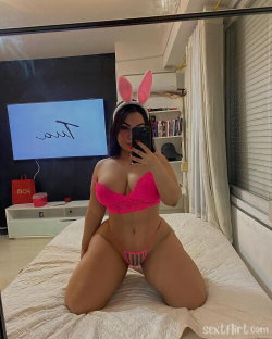 A woman with dark hair wearing pink bunny ears and a matching bright pink lingerie set poses on a bed in a mirror