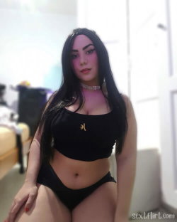 A woman with long dark hair and a curvy build wears a black crop top and matching underwear, sitting indoors near a bed.