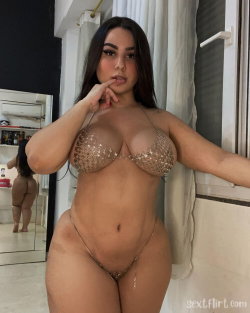 Curvy woman with long dark hair poses in a sparkly gold bra and matching thong, standing near a window with a mirror